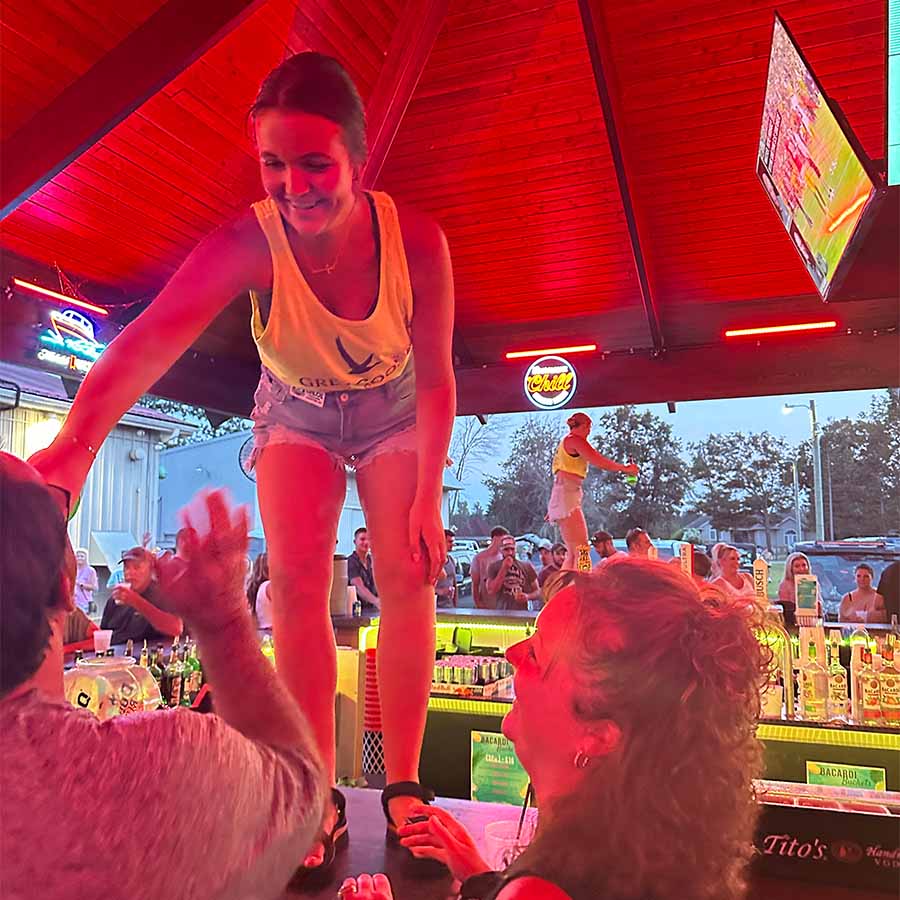 Woman in a yellow tank top dances atop The Dirty Turtle Bar, smiling and reaching for drinks below.