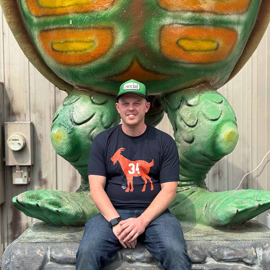 Man in a black t-shirt and green hat sits beneath The Dirty Turtle Bar statue outdoors.