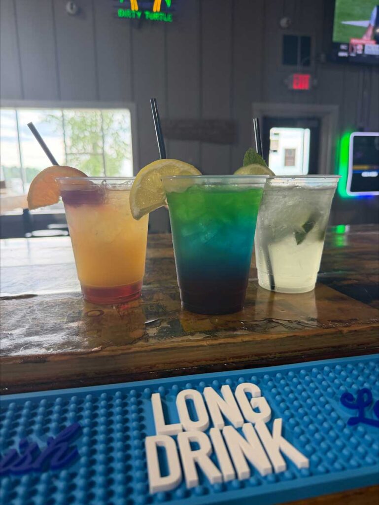 Three colorful cocktails with fruit garnishes on The Dirty Turtle Bar bar, LONG DRINK mat in front.