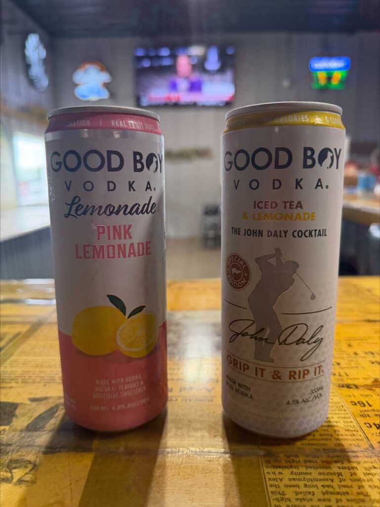 Two cans of Good Boy Vodka sit at The Dirty Turtle Bar with drinks and a TV blurred in the background.