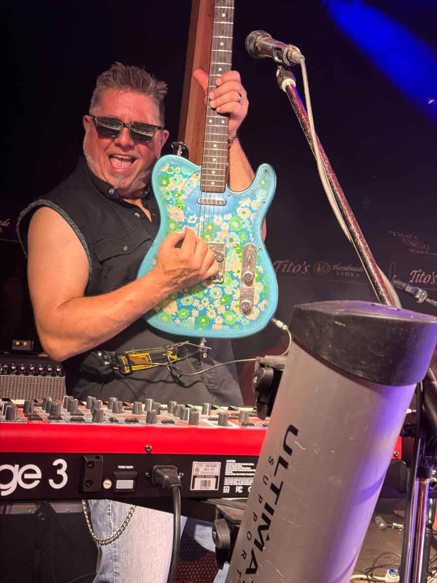 Man in sunglasses plays a blue floral guitar at The Dirty Turtle Bar, smiling energetically on stage.