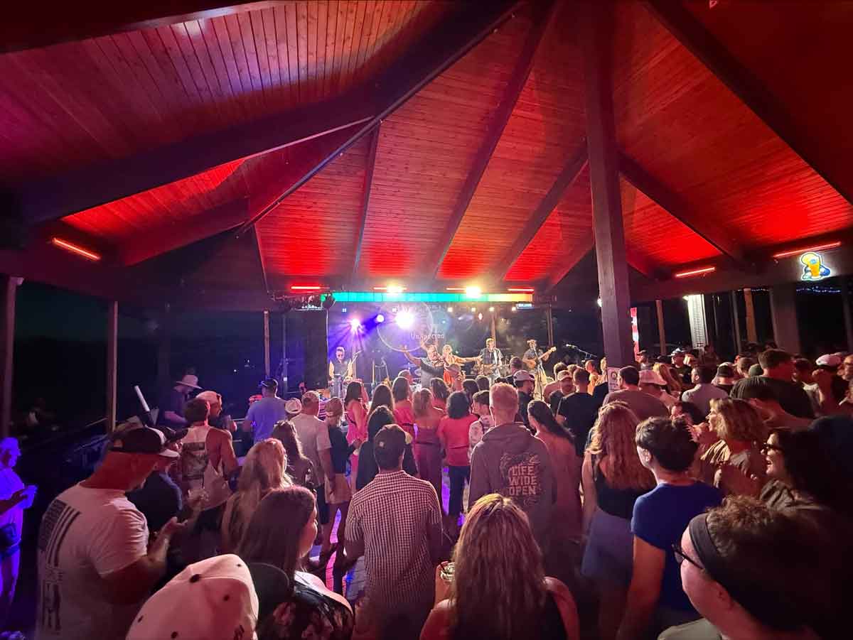 A crowd at The Dirty Turtle Bar enjoys live music, drinks, and colorful lights under a wooden pavilion.