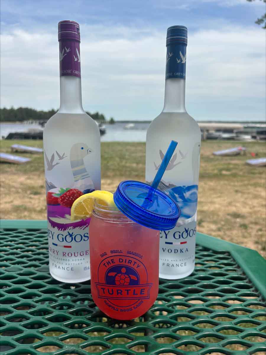 Two vodka bottles and a pink drink from The Dirty Turtle Bar on a picnic table by the lake.