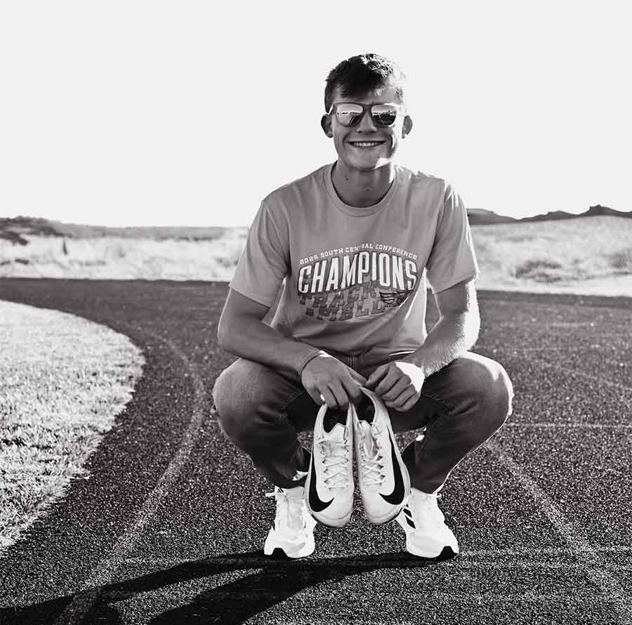 Smiling person in sunglasses squats on a track, holding running shoes, repping The Dirty Turtle Bar.